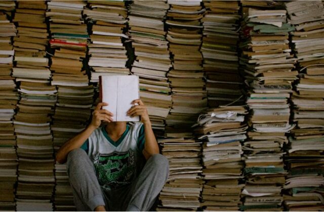 Jakie są najchętniej wybierane lektury szkolne w Polsce? Na zdjęciu chłopak czyta książkę w otoczeniu stosów książek.