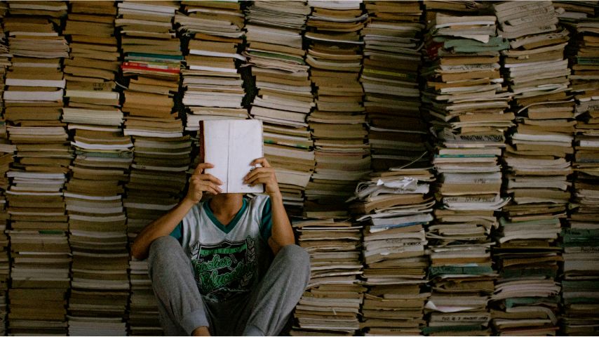 Jakie są najchętniej wybierane lektury szkolne w Polsce? Na zdjęciu chłopak czyta książkę w otoczeniu stosów książek.